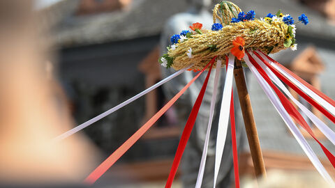 Eine Erntekrone mit bunten Bändern, im Hintergrund ist eine Statue und ein Gebäude zu erkennen.