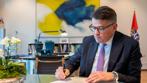 Ministerpräsident Rhein sitzt an einem Glastisch und schreibt etwas auf ein DinA4 Papier.