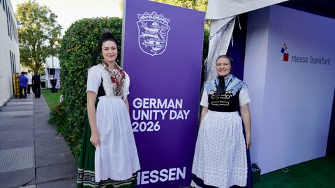 Zwei Frauen in Tracht stehen neben einem Aufsteller "German Unity Day 2026"